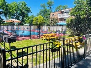 A fenced pool area with lounge chairs and a table, surrounded by greenery and a building in the background.