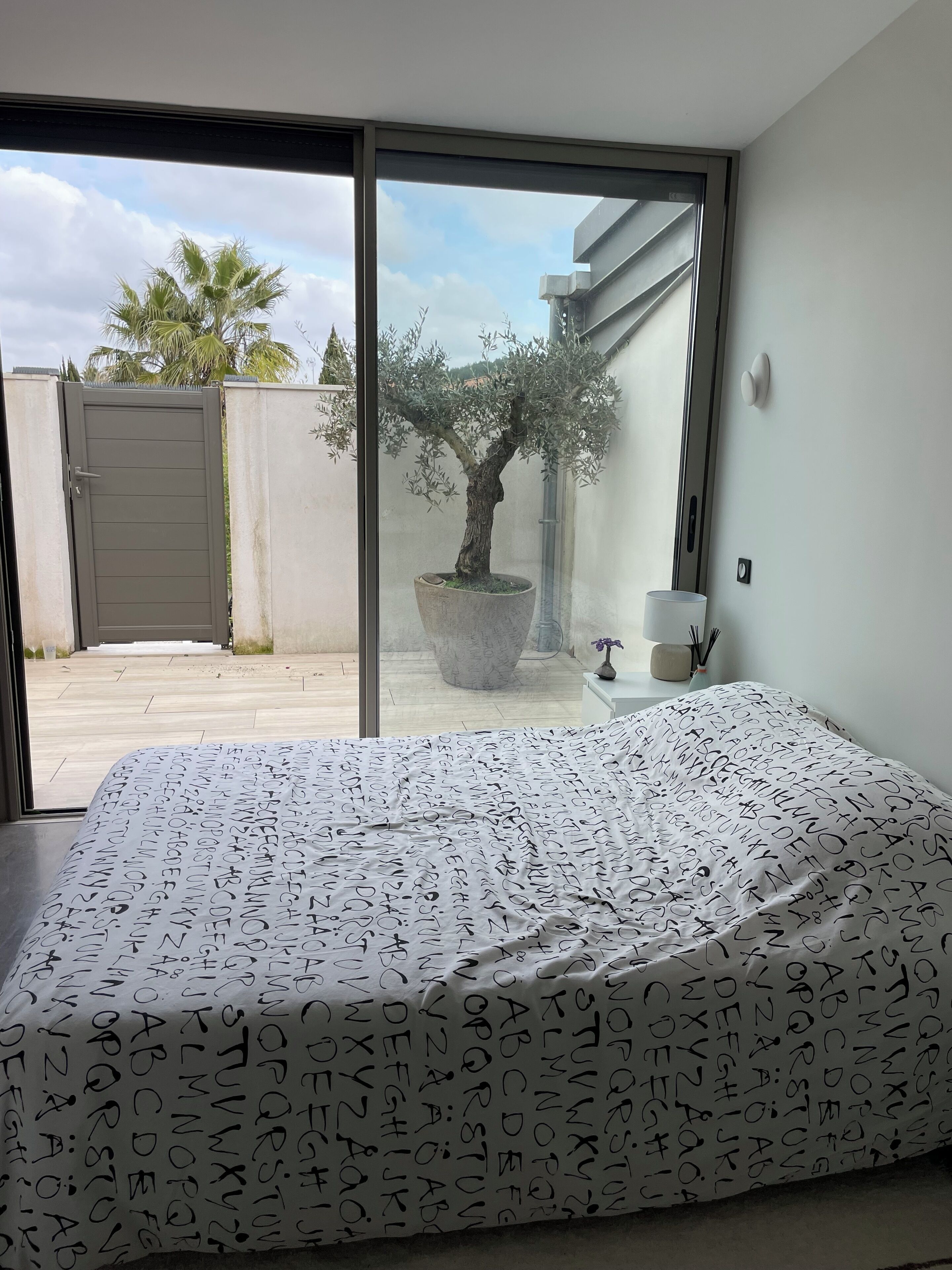 A bedroom with a bed covered in alphabet-patterned bedding, a large window with a view of a palm tree and a gate, and a potted olive tree.
