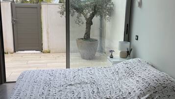 A bedroom with a bed covered in alphabet-patterned bedding, a large window with a view of a palm tree and a gate, and a potted olive tree.