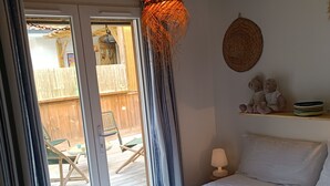 Une chambre avec un lit, un luminaire suspendu, une décoration murale en osier, une petite étagère avec quelques objets, et une vue sur une terrasse extérieure.