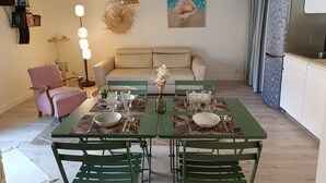 Un coin repas avec une table verte dressée pour quatre personnes, entourée de chaises vertes. Un canapé beige est adossé au mur, et une cuisine avec un réfrigérateur est visible sur la droite.