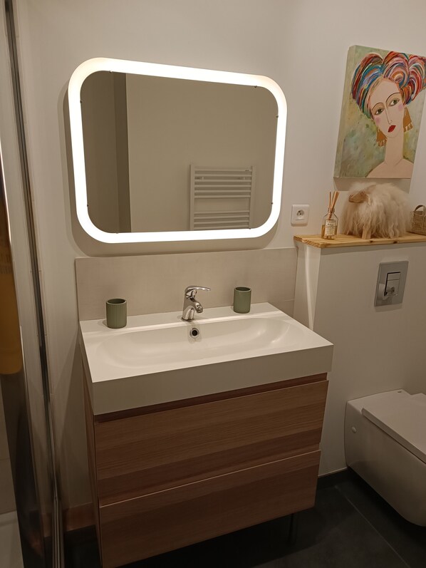 A modern bathroom with a white sink, a mirror with a lighted frame, a towel rack, and a painting on the wall. - La BISCABANE quiet bicycle path near lakes shopping center Classified **** (Biscarrosse)