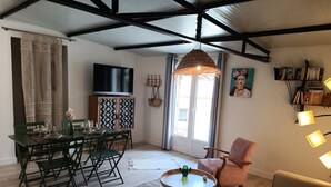 Un salon avec un plafond haut, un coin repas avec une table et des chaises, un canapé, une petite table ronde avec un vase et une bibliothèque.