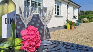 En flaske vin, to vinglas og en lyserød blomst på et bord foran et hvidt hus med vinduer og have.