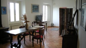 Un coin repas comprenant une table en bois, des chaises, un vase avec des fleurs, un téléviseur et une fenêtre avec des stores.