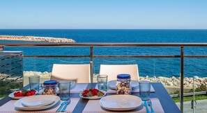 Una mesa para dos personas, con platos, cubiertos y copas, con vistas a una costa rocosa y el mar.