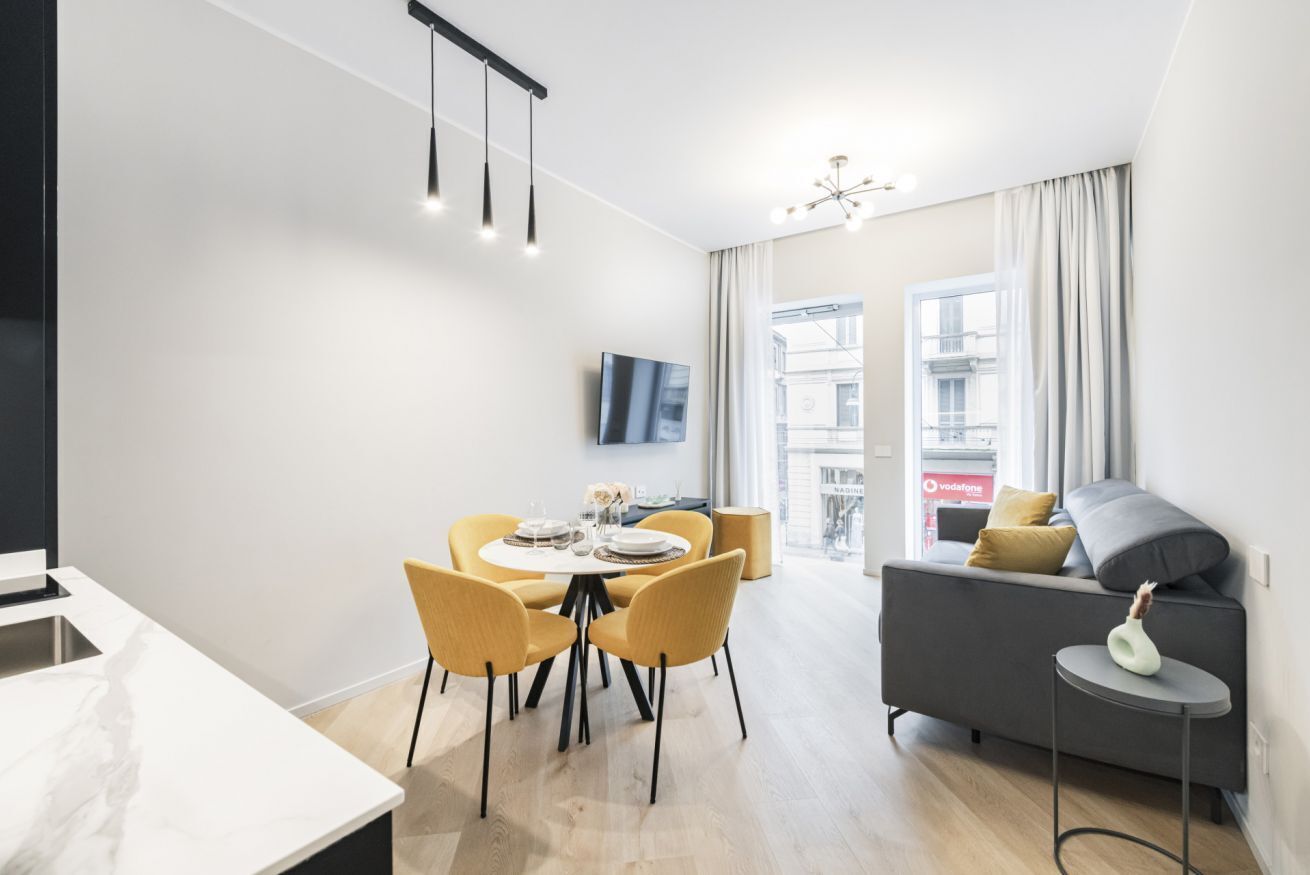 Un salon moderne comprenant un canapé, une table à manger avec des chaises, un téléviseur fixé au mur et une vue sur l’extérieur grâce à de grandes fenêtres.