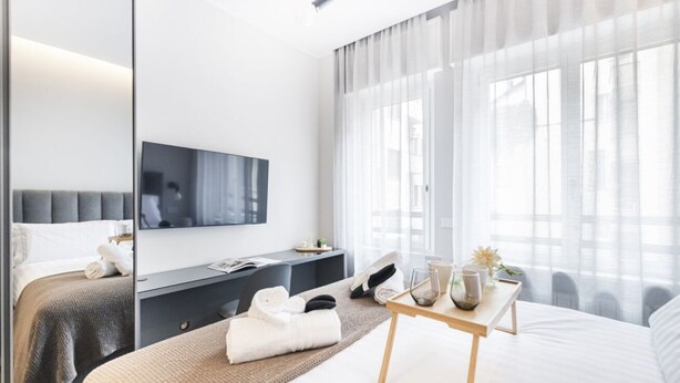 Una habitación de hotel moderna con una cama grande, una mesita de noche, un televisor de pantalla plana y un ventanal con cortinas traslúcidas.