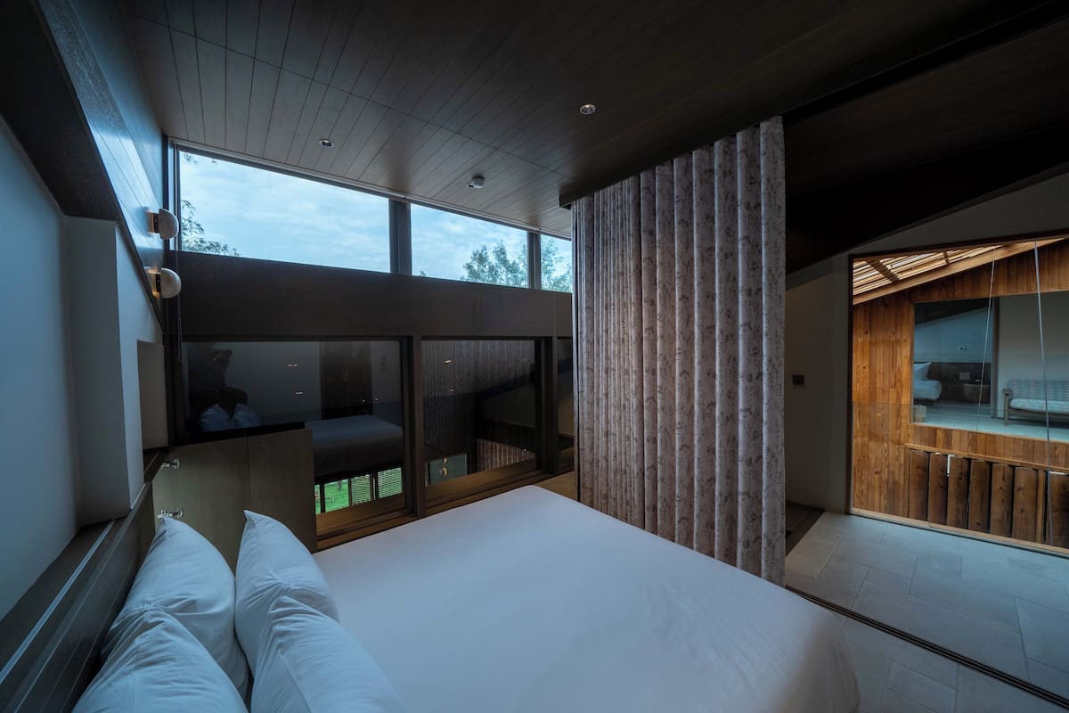 A modern bedroom with a large bed, a glass partition, and a view of the outdoors.