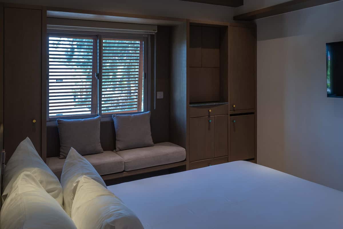 A bedroom with a bed, a window with blinds, a built-in wardrobe, and a flat-screen TV mounted on the wall.