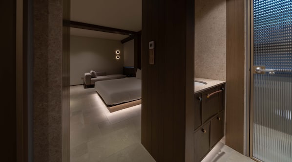 A modern bedroom with a large bed, a bedside table, and a nightstand. There is a bathroom with a sink and a shower.