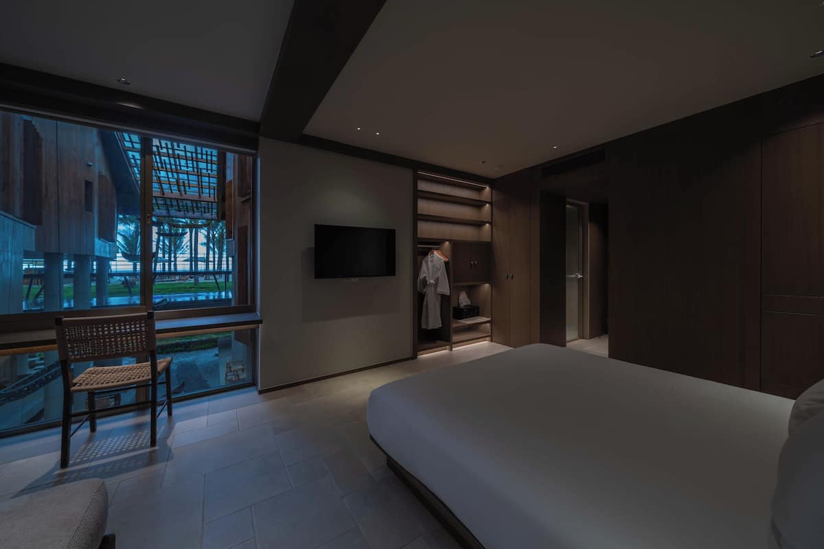 A modern bedroom with a large bed, a TV mounted on the wall, and a window with a view of a building.