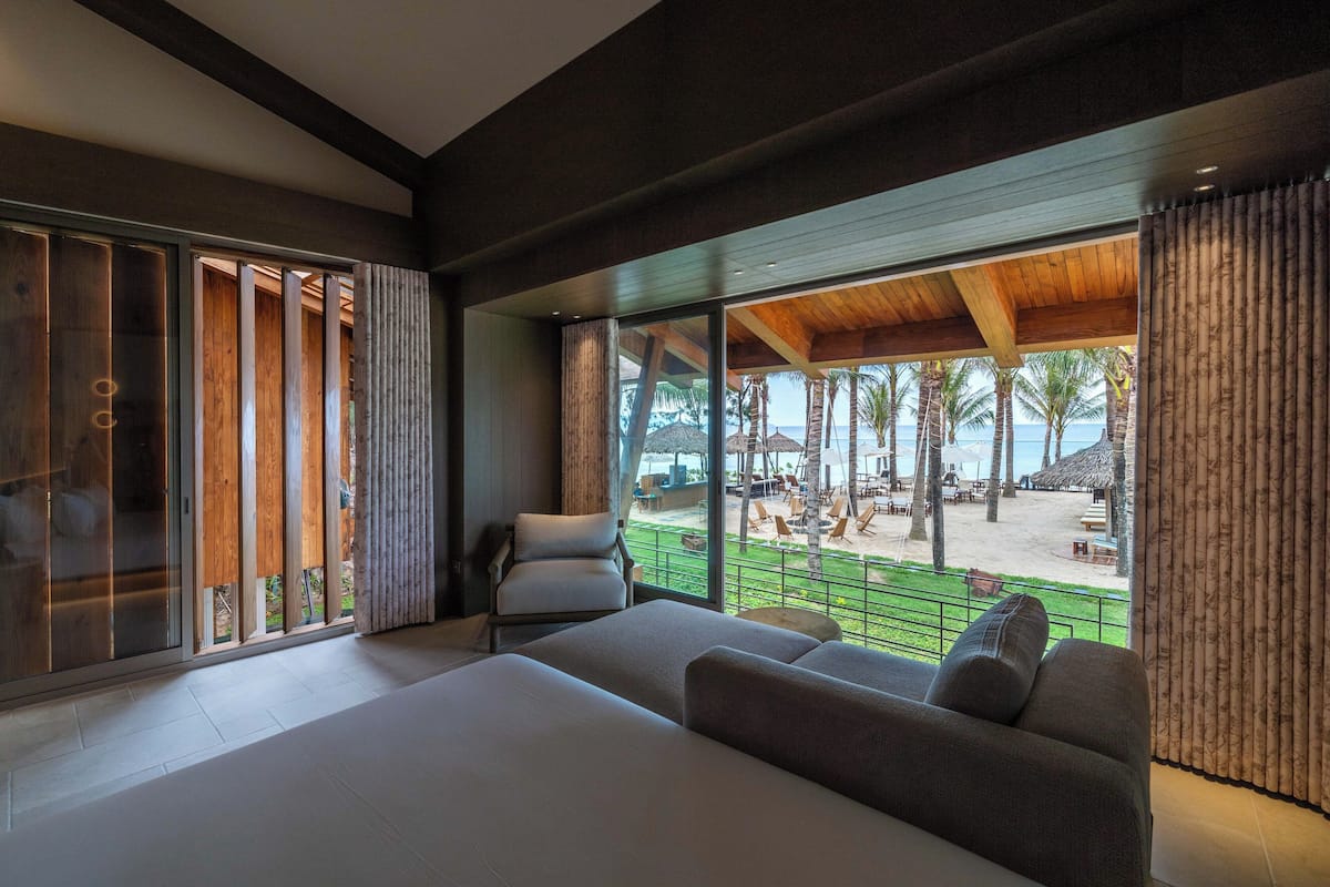 A modern hotel room with a large bed, a seating area, and a view of a beach and palm trees.