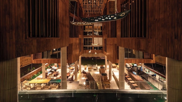Un vaste atrium doté d’un haut plafond et de poutres en bois, avec un espace central ouvert comprenant des sièges et des tables, entouré de plusieurs niveaux de balcons et offrant une vue sur l’étage inférieur.