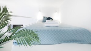 A neatly made bed with a blue patterned bedspread and white pillows, a small bedside table with a lamp, and a potted plant to the left.