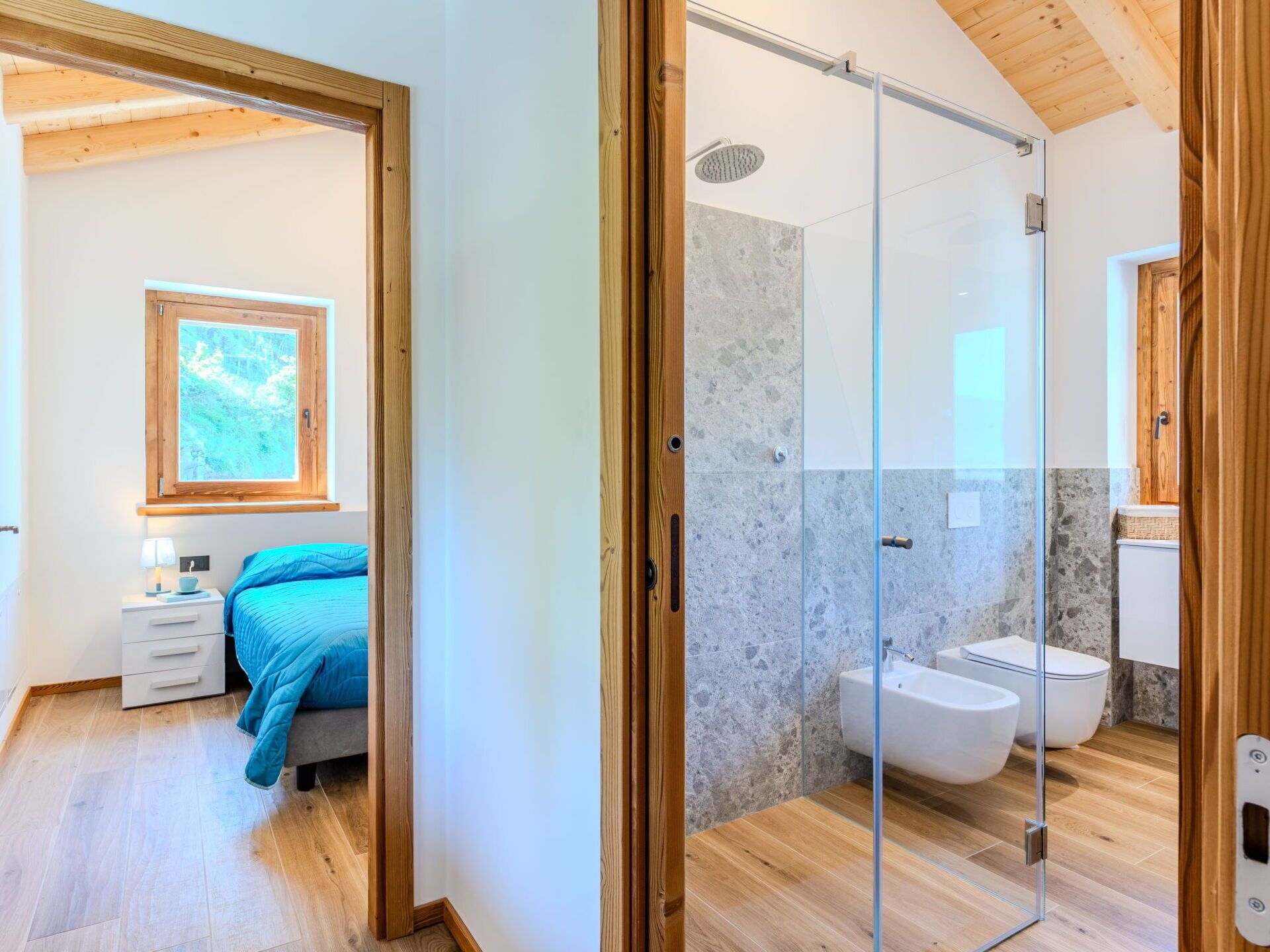 A modern bedroom with a bed, bedside table, and a window. Adjacent is a bathroom with a shower, toilet, and sink.