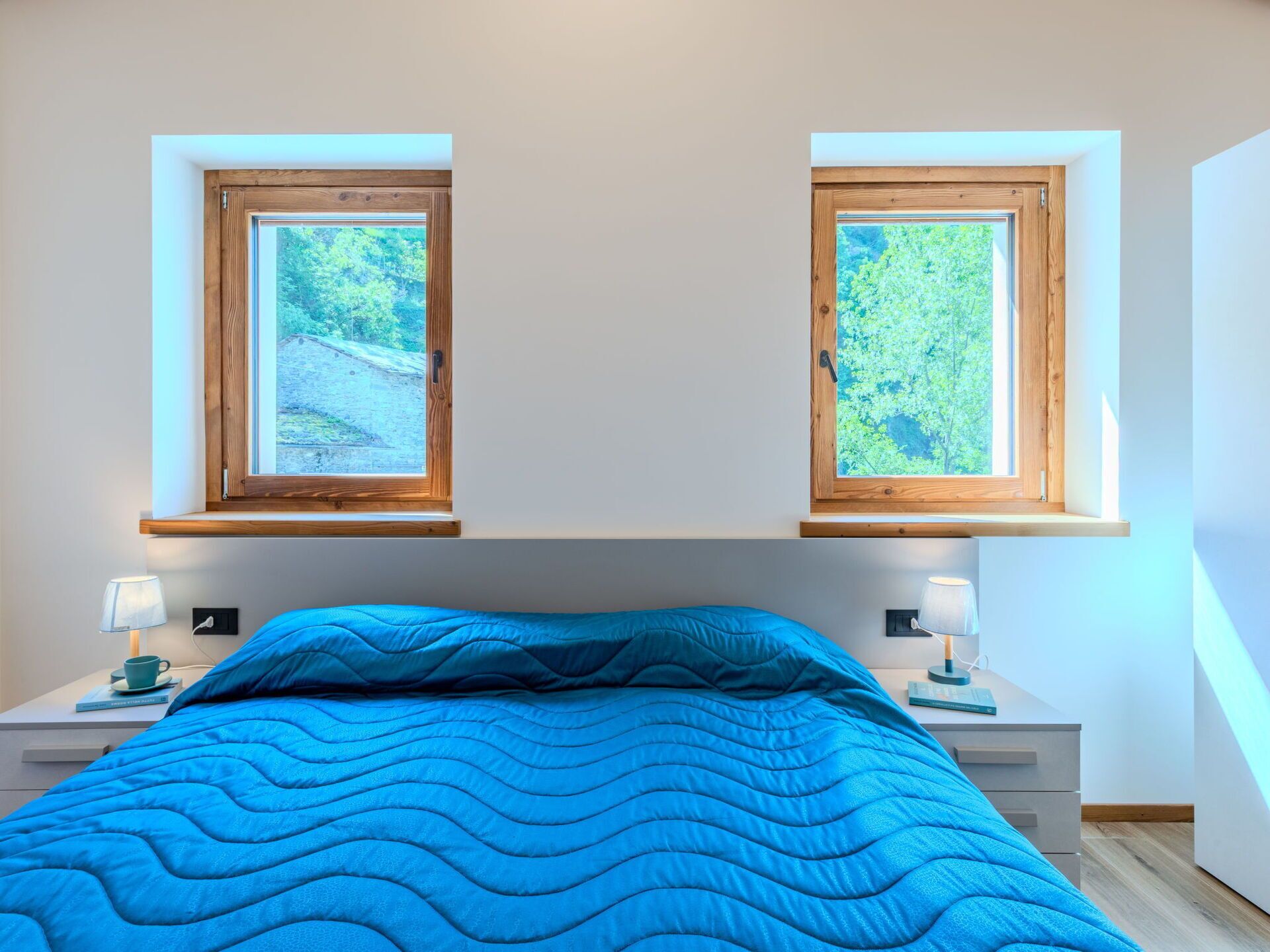 Une chambre à coucher avec un lit, deux tables de chevet, deux fenêtres et une vue sur la verdure.
