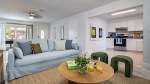 A modern living room with a sofa, a coffee table with a vase and fruit, and a kitchen in the background.