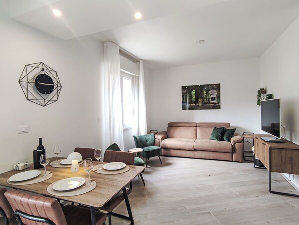 En moderne stue med sofa, spisebord dækket til fire personer, fjernsyn på et træstel og et geometrisk vægur.