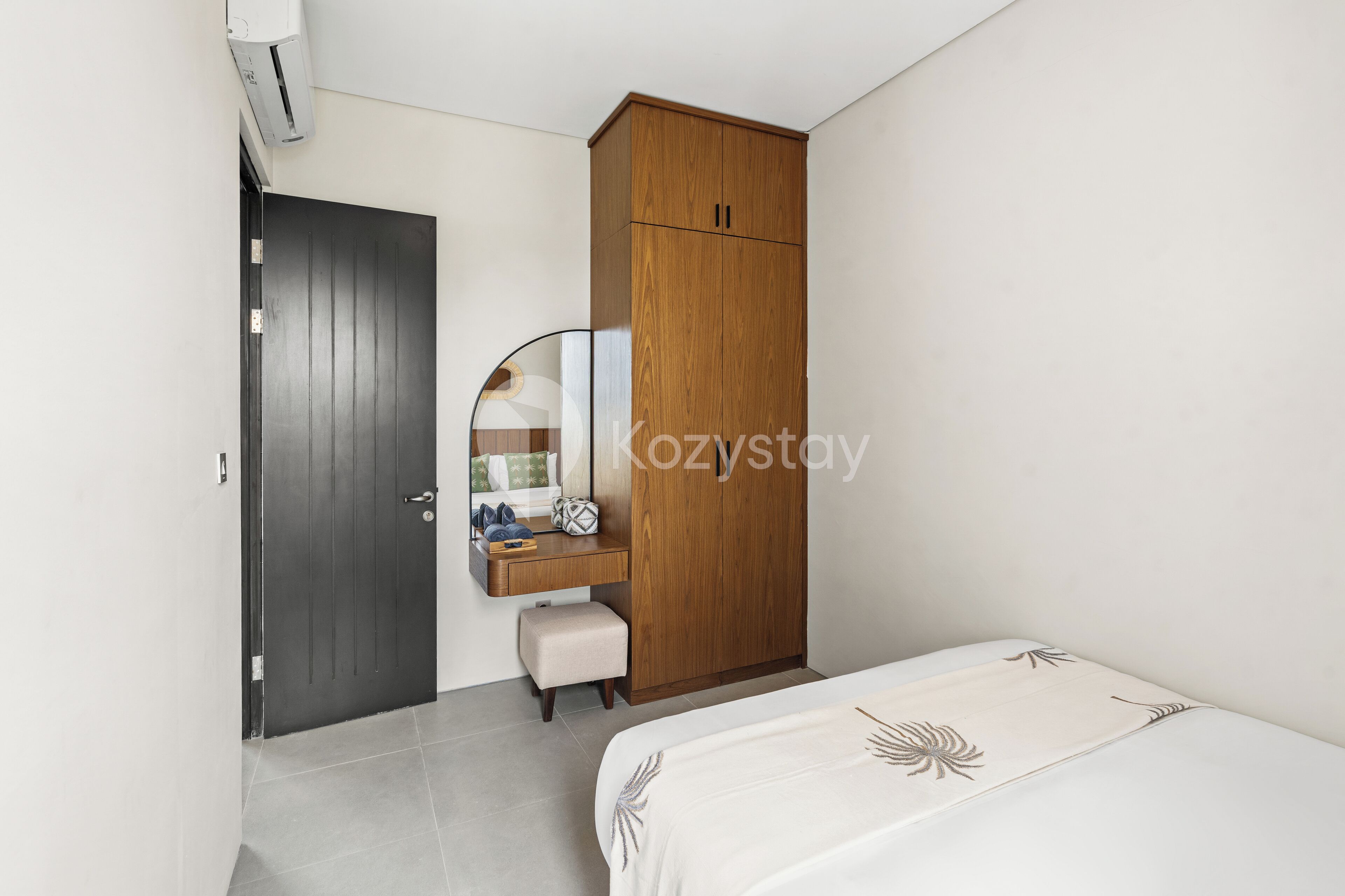 A bedroom with a bed, a wooden wardrobe, a small desk with a mirror, and a door.