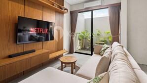 Ein modernes Wohnzimmer mit einem an einer Holzwand montierten Flachbildfernseher, einer Couch, einem Couchtisch und einer Glastür zum Balkon mit Pflanzen.
