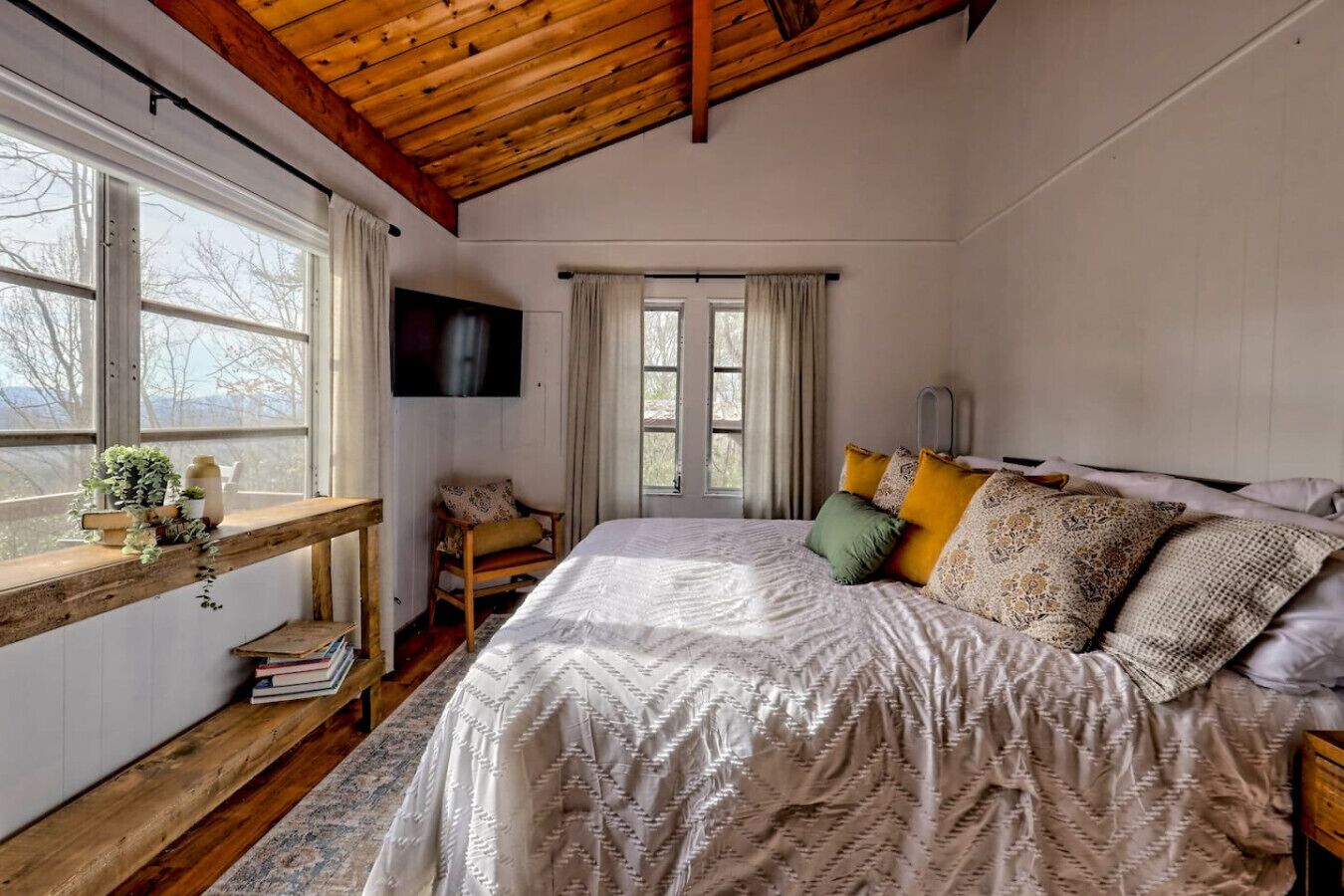 A bedroom with a large bed, wooden headboard, a wooden bench with books and plants, a TV mounted on the wall, and a window with curtains.