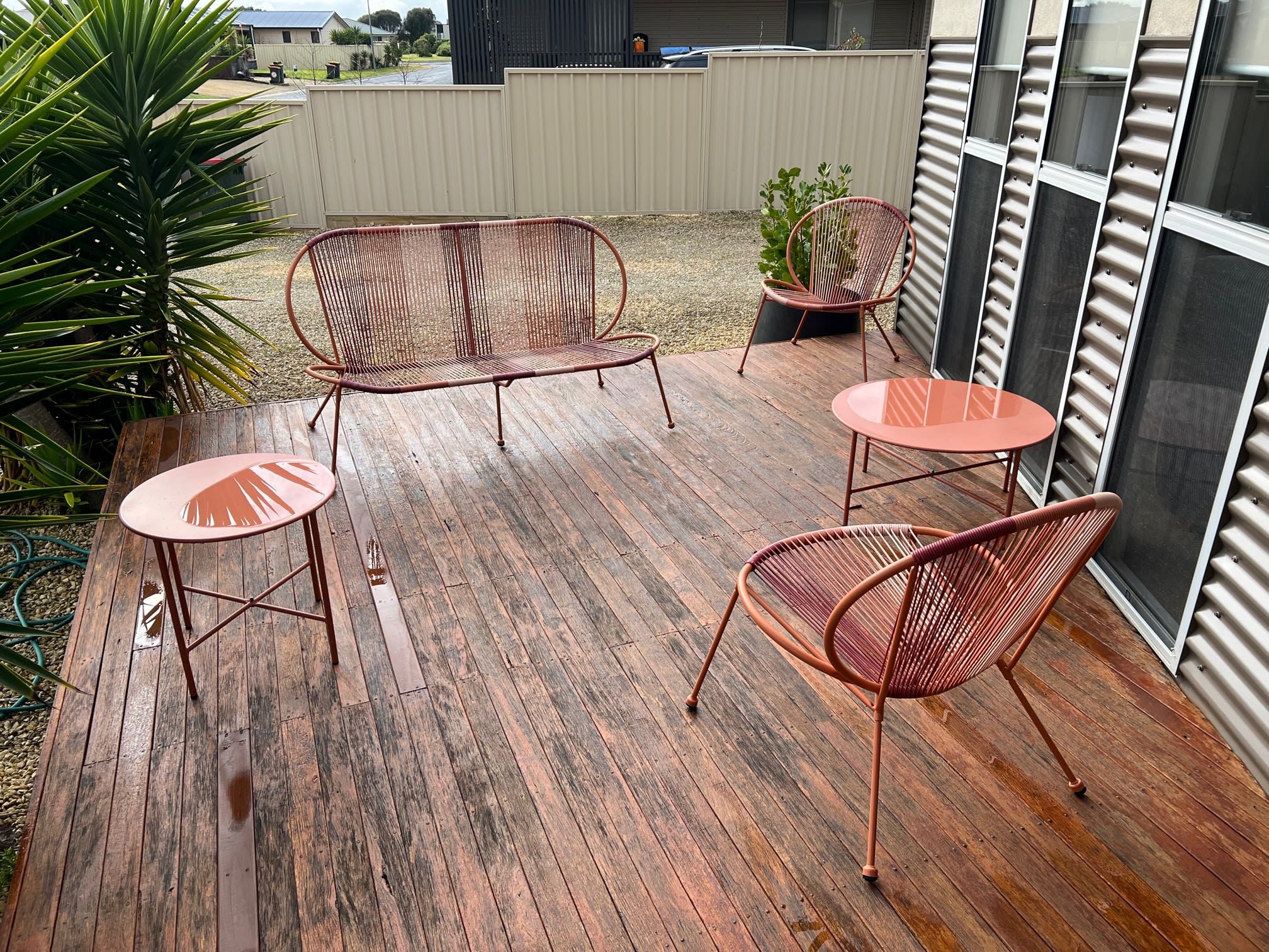 A wooden deck with metal chairs and a table, surrounded by plants and a view of a residential area.