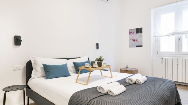 A modern hotel room with a neatly made bed, a small bedside table, a bedside lamp, and a window with curtains. - EASYLIFE – Your ideal space in Milan (Milano)