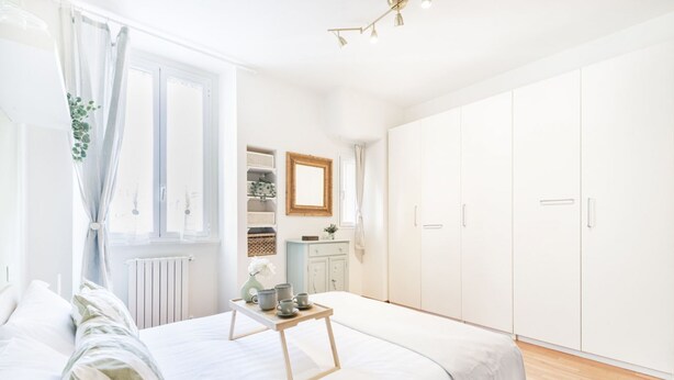 Un dormitorio con cama, mesita de noche con tazas y un florero, armario y ventana con cortinas.