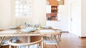 Comedor con mesa de madera dispuesta para cuatro personas, rodeada de sillas con cojines. Se ve un ventanal con vidrio decorativo, y al fondo un área de cocina con armarios blancos y encimera.