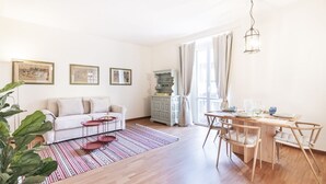 Una sala de estar con sofá, una mesa de comedor con sillas, una mesita de centro, un tapete estampado y un armario.