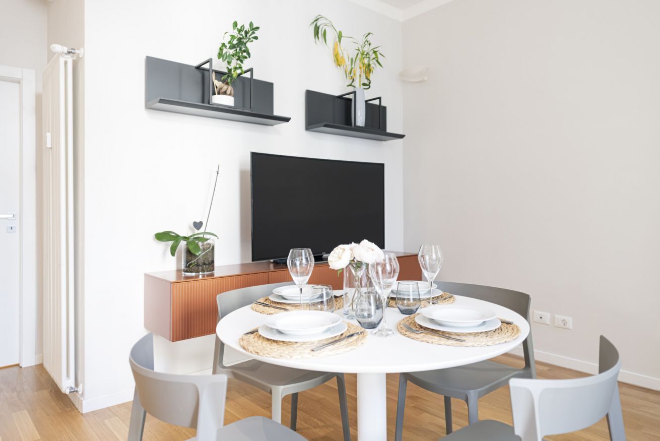 A modern dining area with a round table set for four, surrounded by grey chairs. There are two black shelves with plants on the wall above the table.