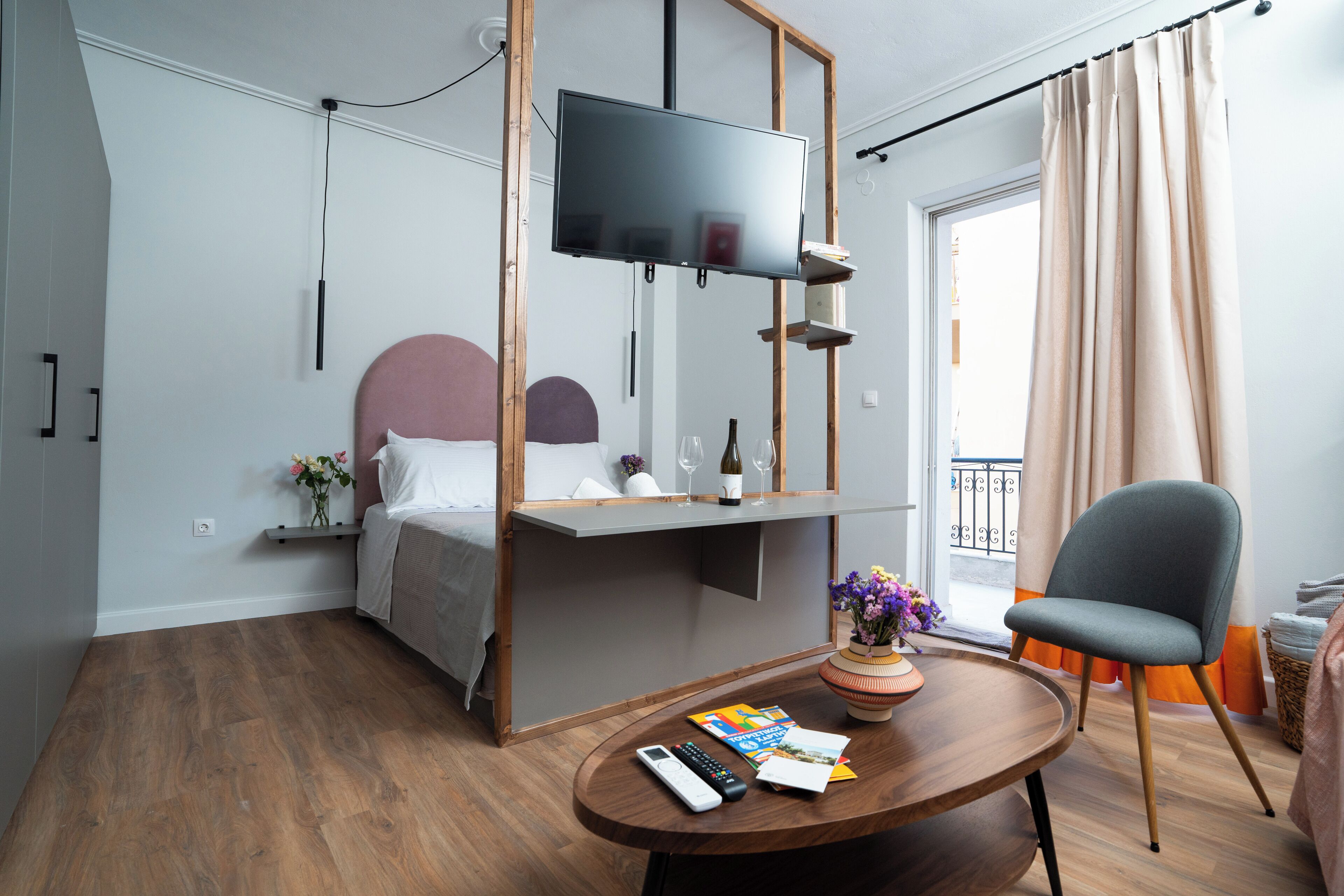 A modern hotel room with a bed, a mounted TV, a small table with a remote control, a chair, and a window with curtains.