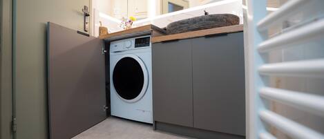 A compact laundry room with a washing machine, a drying rack, and a small shelf with a basket and flowers.