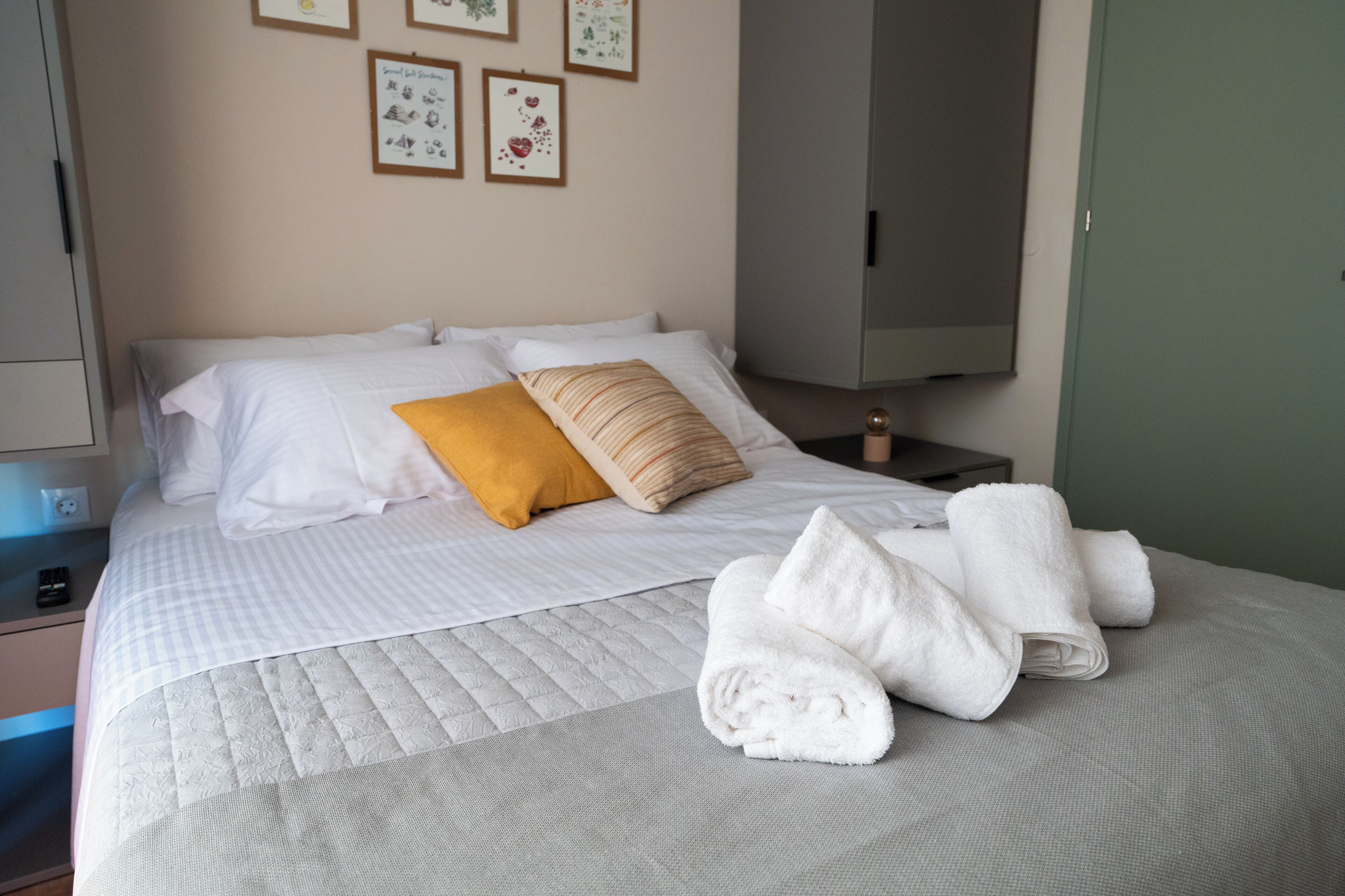 A neatly made bed with white and yellow pillows, a folded white towel, and a bedside table with a lamp and a small decorative item.