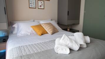 A neatly made bed with white and yellow pillows, a folded white towel, and a bedside table with a lamp and a small decorative item.