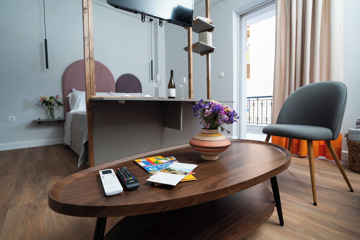 Ein modernes Hotelzimmer mit einem Holztisch, einem Bett mit Kopfteil, einem kleinen Beistelltisch mit Blumen und einem grauen Sessel.