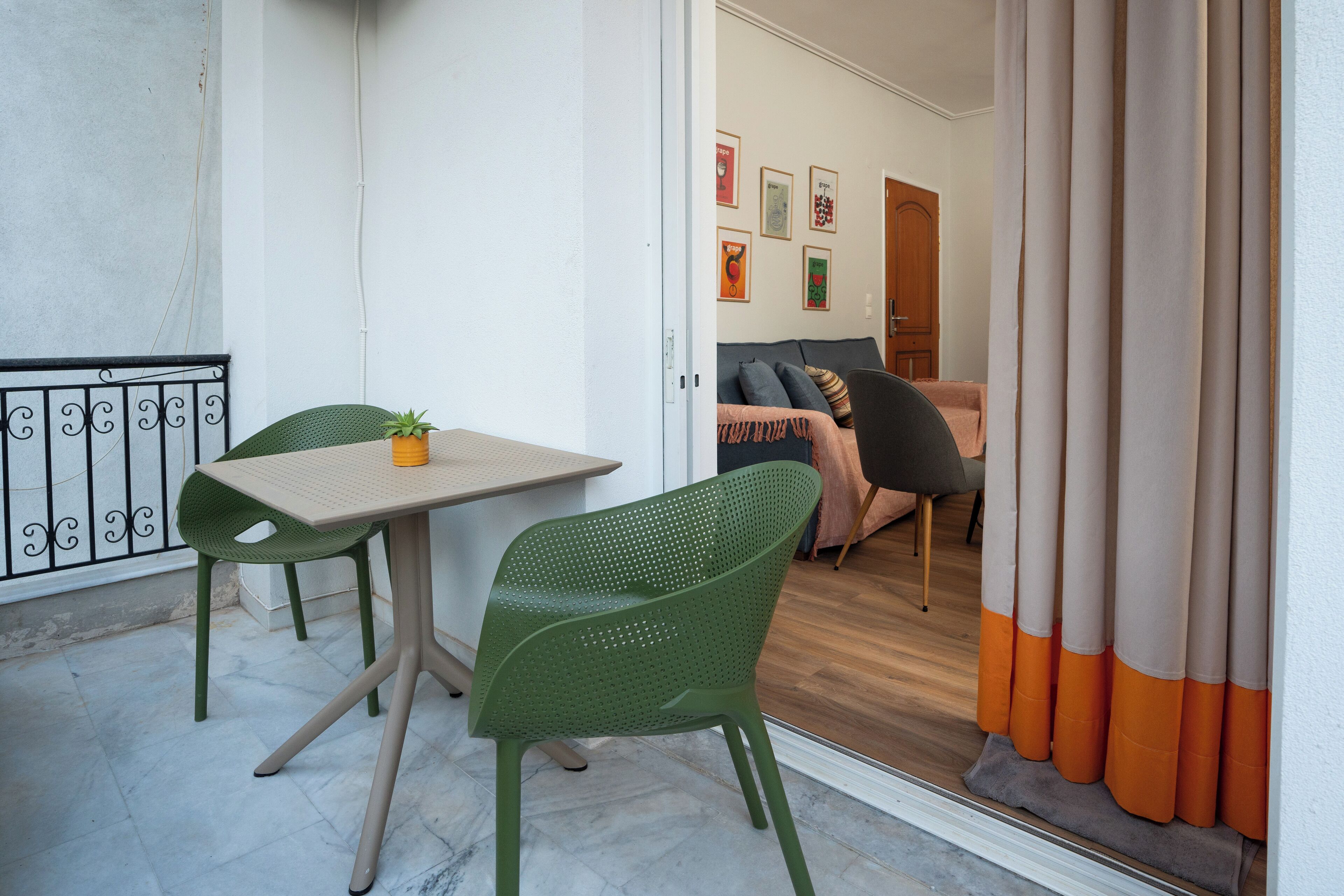 A balcony with a table and chairs, a potted plant, and a view into an interior space with a sofa and curtains.