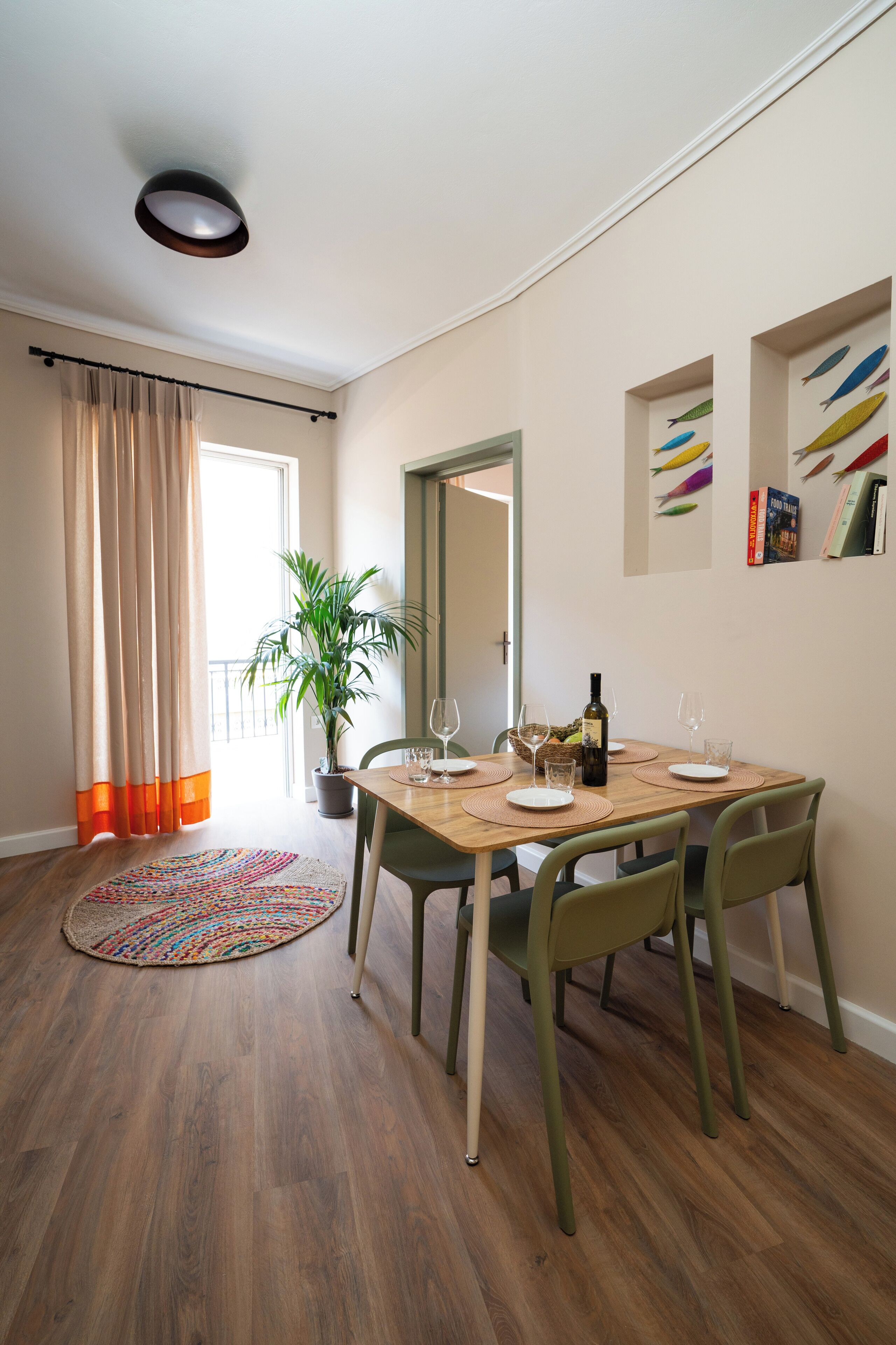 Un comedor con una mesa de madera dispuesta para cuatro, una planta en maceta y vistas del balcón a través de una ventana con cortinas color beige.