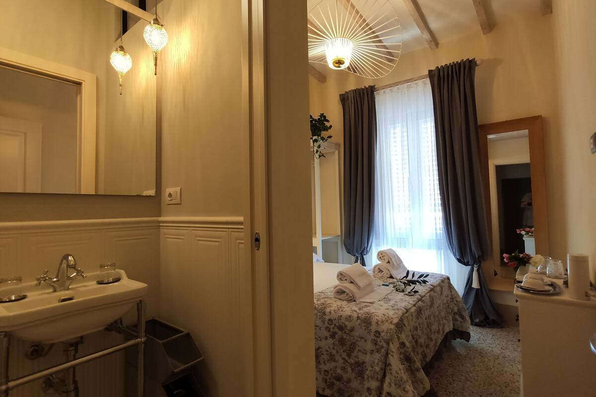 A hotel room with a bathroom featuring a sink, mirror, and hanging lights. The room has a bed with a floral bedspread, a bedside table, and a window with curtains.