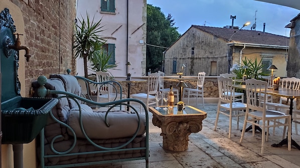 Un'area esterna con tavoli in pietra e sedie bianche, circondata da muri in pietra e piante in vaso.