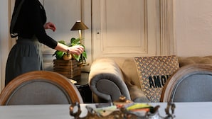 A woman standing by a table with a decorative tray, a lamp, and a cushion with a pattern. - Residenza d'Epoca CAVALIERE (Bibbona)