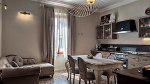 A cozy living room with a sofa, a dining table with chairs, a kitchen area with cabinets and appliances, and a large window with curtains. - Residenza d'Epoca CAVALIERE (Bibbona)