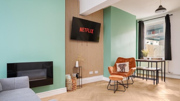 A modern living room with a flat-screen TV mounted on a wall, a grey sofa, a brown armchair with a decorative pillow, a small table with a plant, and a window with curtains. - Free Parking | Near Aintree Racecourse & Anfield | Ideal for Contractors & Families (Liverpool)