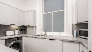 Cocina moderna con muebles blancos, lavadora integrada, un lavabo y un ventanal con vistas decorativas.