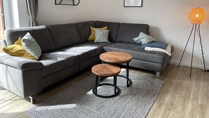 Ein modernes Wohnzimmer mit einem grauen Ecksofa, zwei Holztischen, einem grauen Teppich mit Muster, einer Stehlampe und einem an der Wand befestigten Bücherregal mit Büchern und einem kleinen dekorativen Gegenstand.