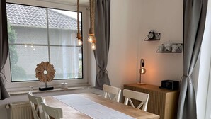 Ein Essbereich mit einem Holztisch, weißen Stühlen, einer Pendelleuchte und einem Fenster mit Vorhängen.