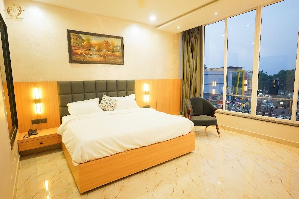 A hotel room with a large bed, a nightstand, a chair, and a city view through large windows.