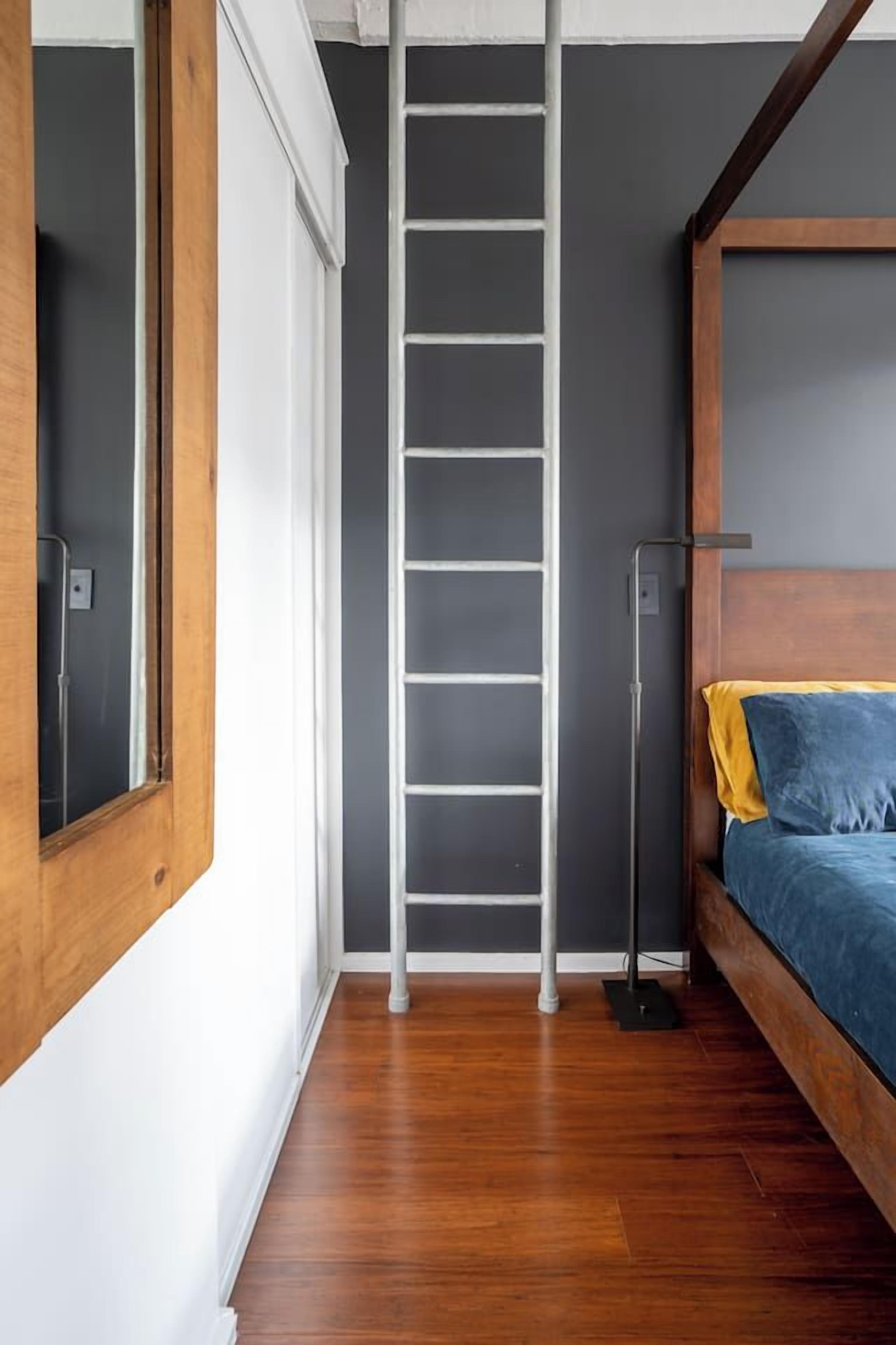 A room with a wooden floor, a bed with blue bedding, a ladder against the wall, and a window with a wooden frame.