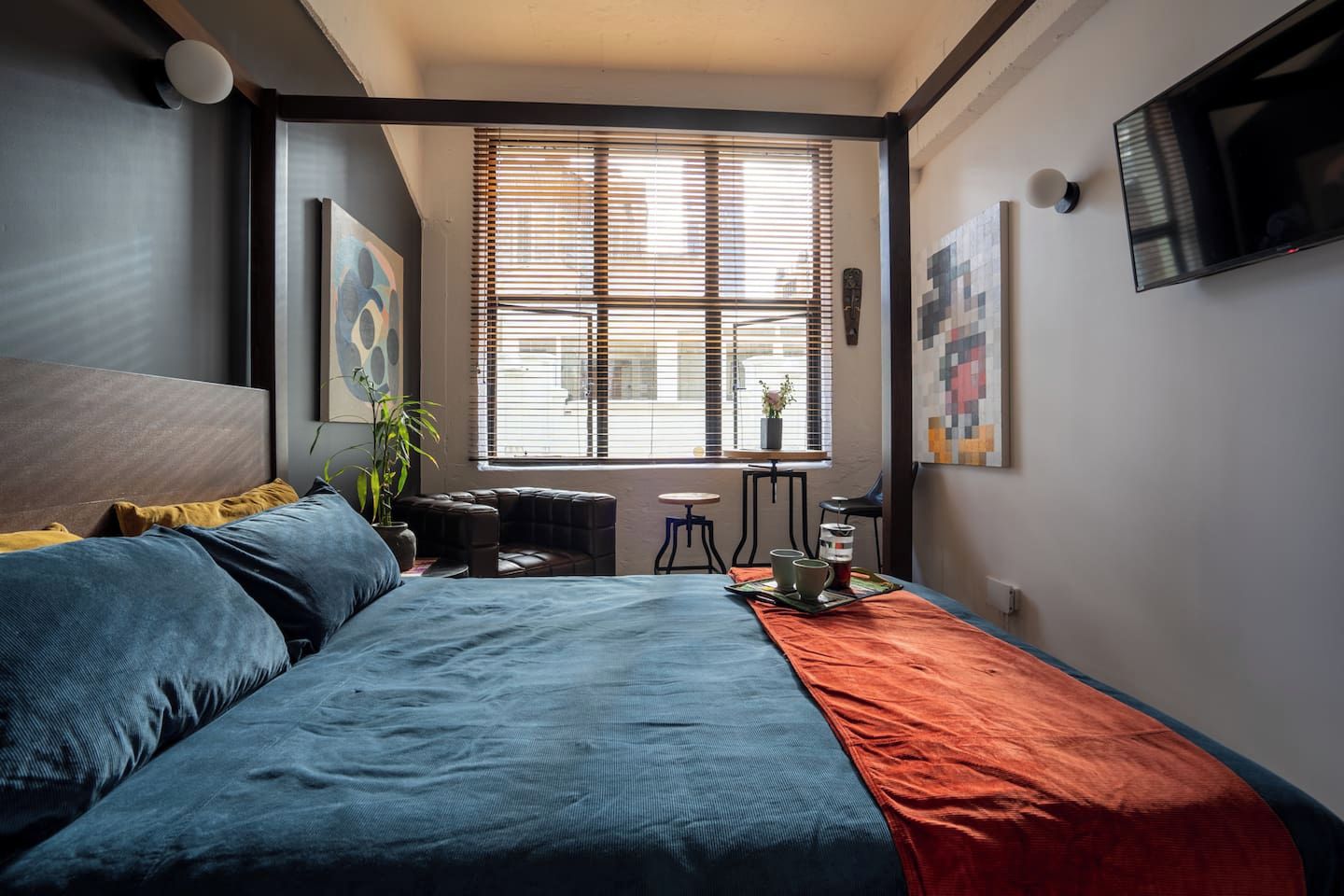 Ein Schlafzimmer mit einem Bett, einem Fenster mit Jalousien, einem kleinen Tisch mit Tassen und einem Fernseher an der Wand.